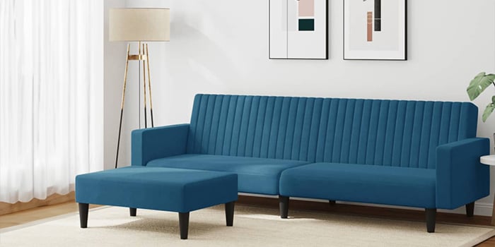Moderni mėlyno aksomo kampinė sofa su vertikaliomis siūlėmis ir derančiu pufu. Sofa turi juodas medines kojeles ir stovi šviesioje svetainėje su įrėmintais paveikslais ant sienos bei toršeru kampe.